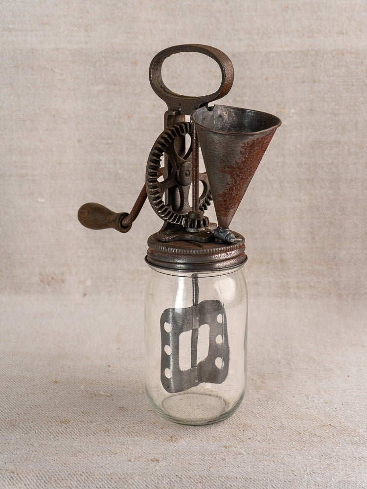 Antique French mayonnaise maker, c. 1900, with cast iron crank and glass jar, on fabric backdrop.