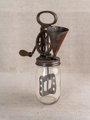 Antique French mayonnaise maker, c. 1900, with cast iron crank and glass jar, on fabric backdrop.