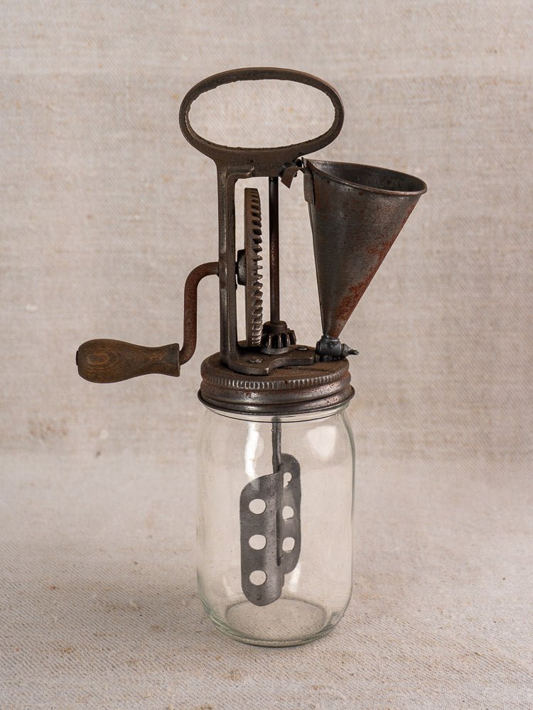 Antique French mayonnaise maker with cast iron crank and glass jar, displayed on neutral textured fabric.