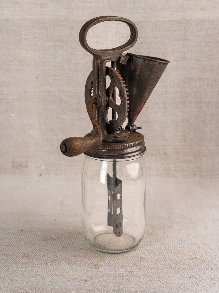 Antique French mayonnaise maker with cast iron hand crank on lidded glass jar, neutral fabric backdrop.