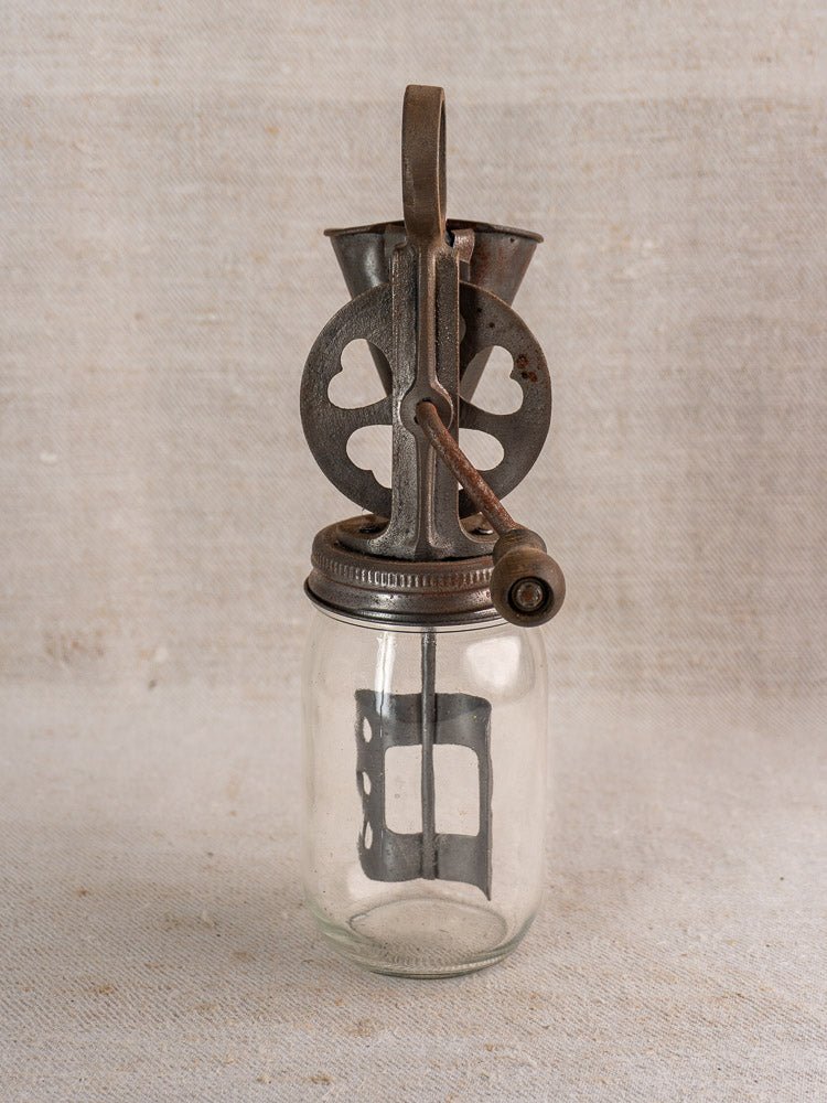 Antique French cast iron hand-crank egg beater with heart motif on clear glass jar, neutral fabric background.