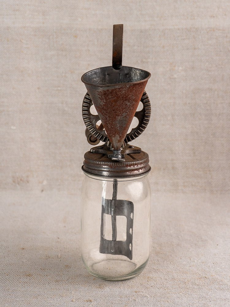 Antique French mayonnaise maker with cast iron crank, glass jar, metal lid, and gears on fabric.