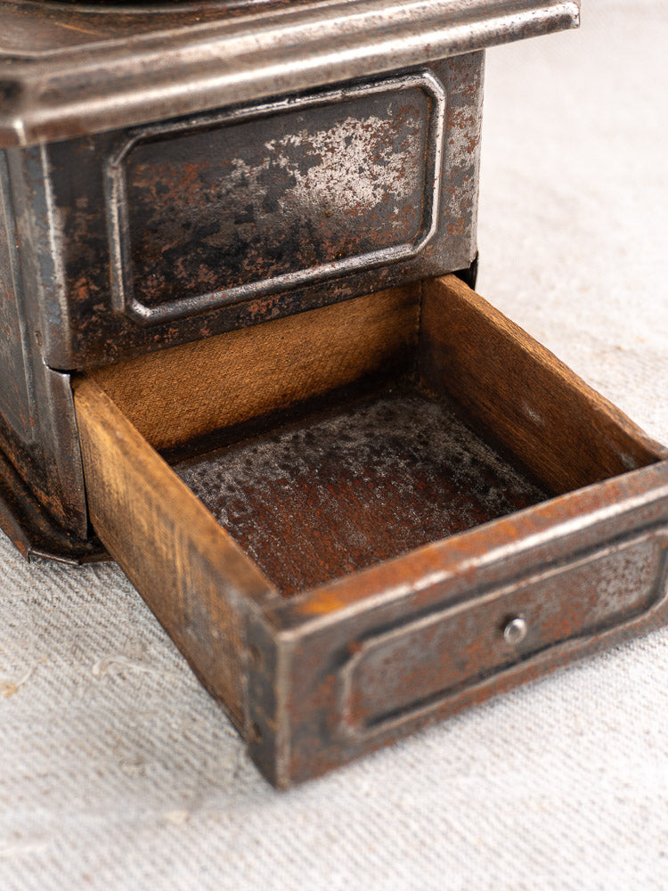 Antique French Peugeot Frères tinned steel coffee mill with open wooden drawer on light fabric.