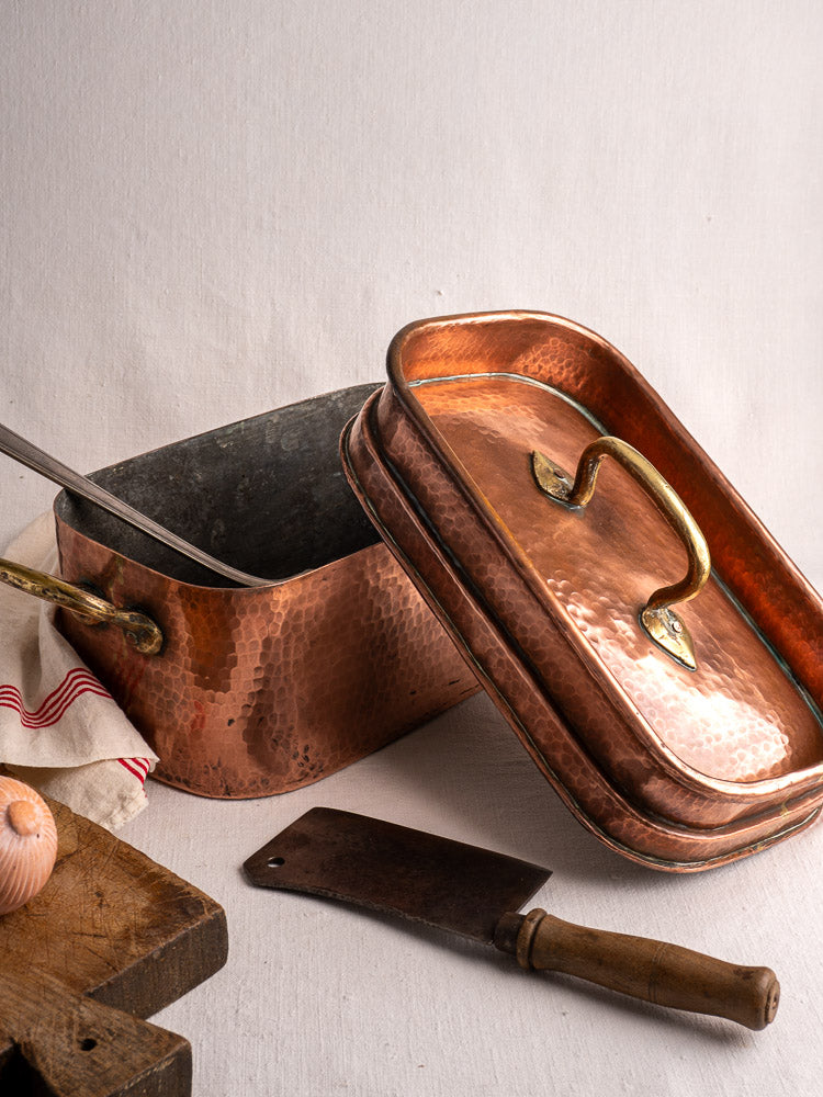 Antique French copper daubière with brass handle and lid, metal ladle inside; onion and cleaver rest on wooden board nearby.