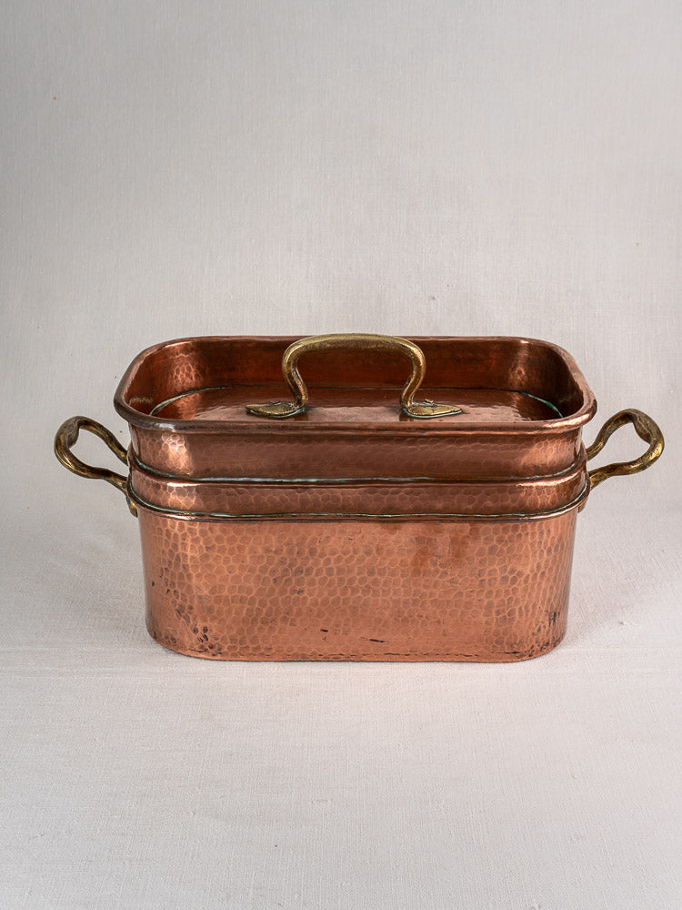 Antique French copper daubière with lid and brass handles, hand-hammered, on a light surface against a neutral background.