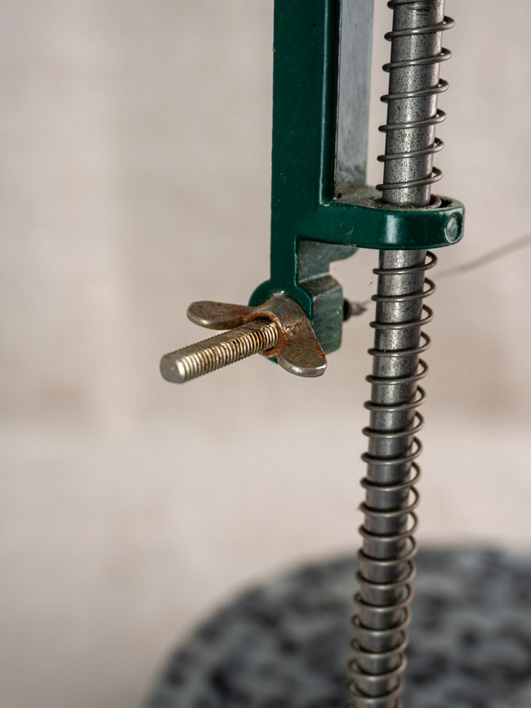Metal spring, threaded bolt, and wing nut detail on green frame of 1960s French Roquefort Cheese Cutter; blurred background.