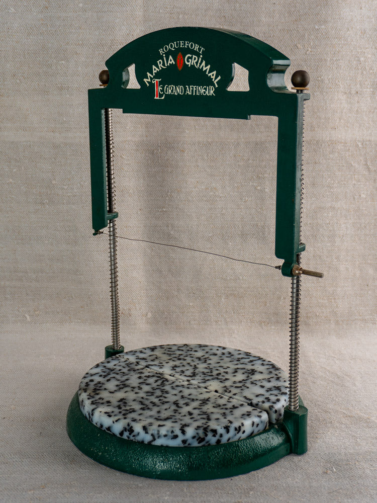 Vintage French Roquefort cheese cutter by Maria Grimal Fromagerie slicing speckled cheese wheel on neutral fabric background.