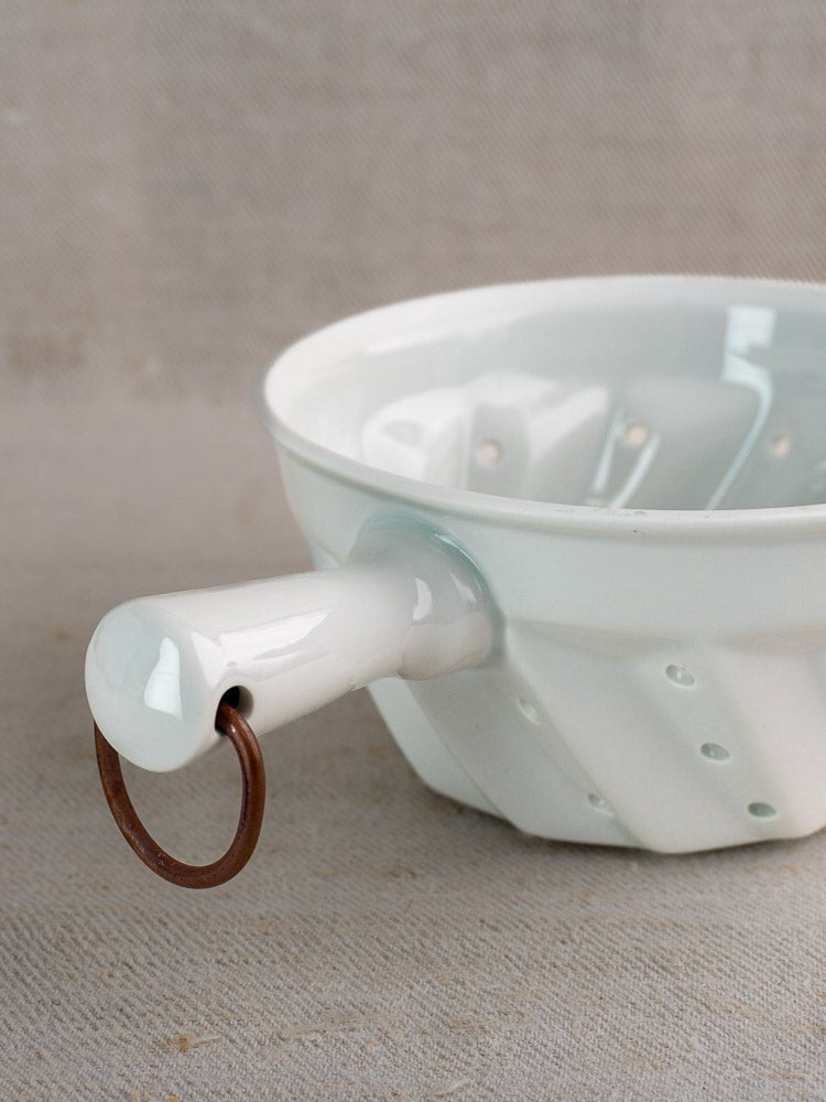 French vintage white porcelain colander with handle, copper ring, ribbed design on beige surface.