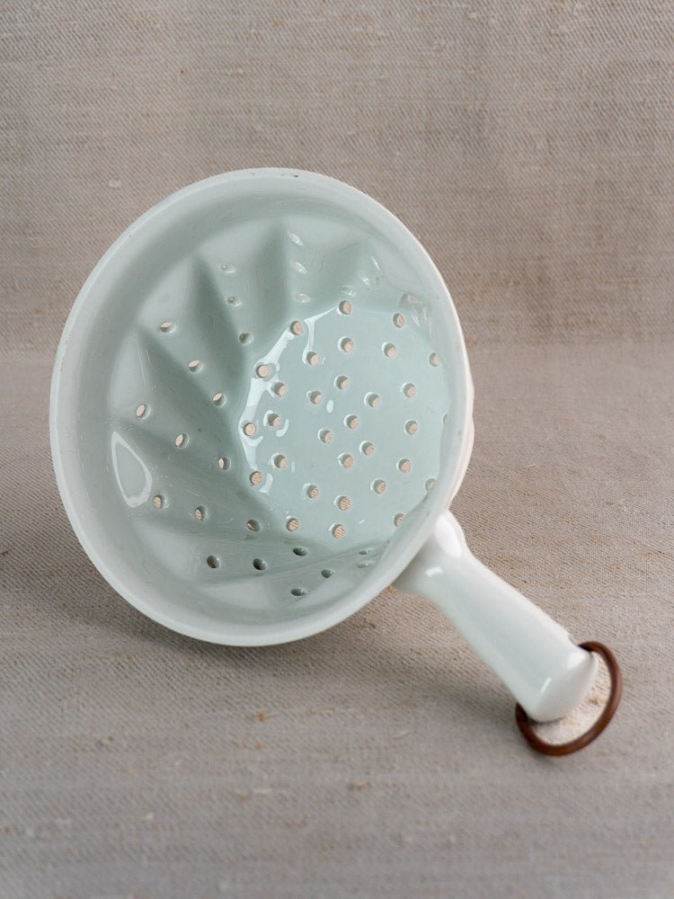 French porcelain berry strainer with white enamel finish, sturdy handle, and copper ring on beige fabric.