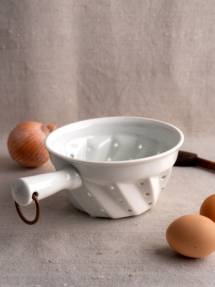 White ceramic berry strainer with copper ring handle, inspired by vintage French porcelain, on beige fabric.