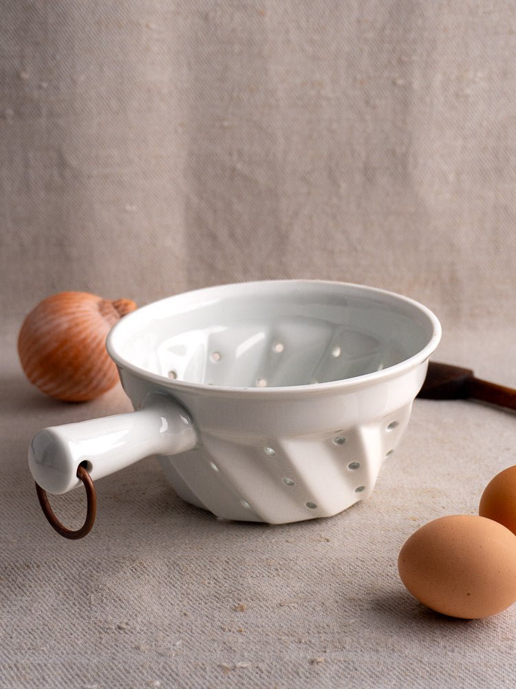 French porcelain berry colander with copper ring on beige fabric, two eggs and onion alongside.