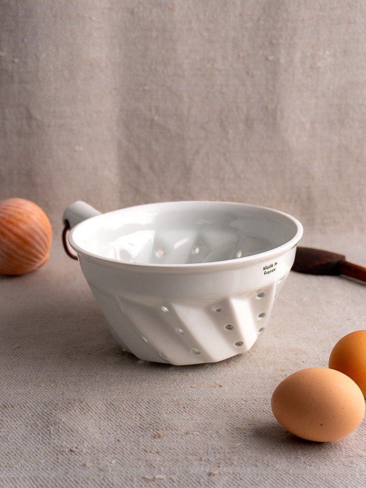 French vintage white porcelain colander with copper ring, displayed on light fabric with eggs and wooden utensil.