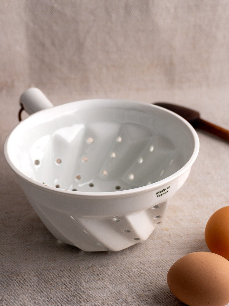 French vintage white porcelain colander with copper ring, on textured surface with eggs and wooden utensil.