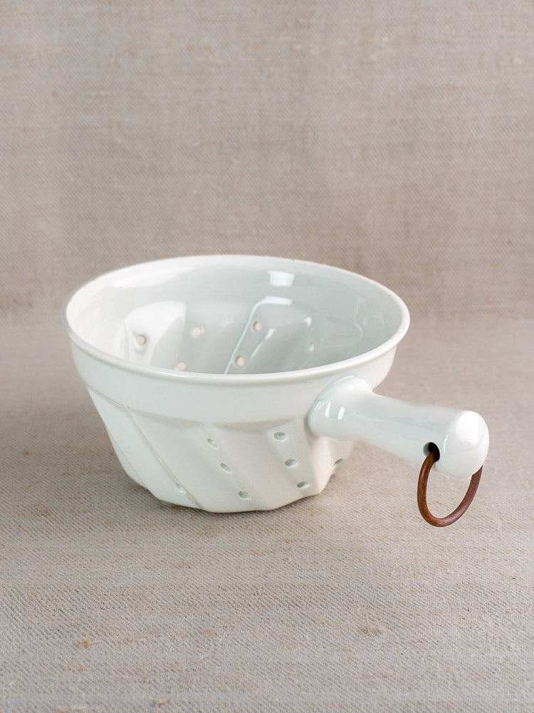 White ceramic berry strainer with copper ring handle, inspired by vintage French porcelain, on beige fabric.