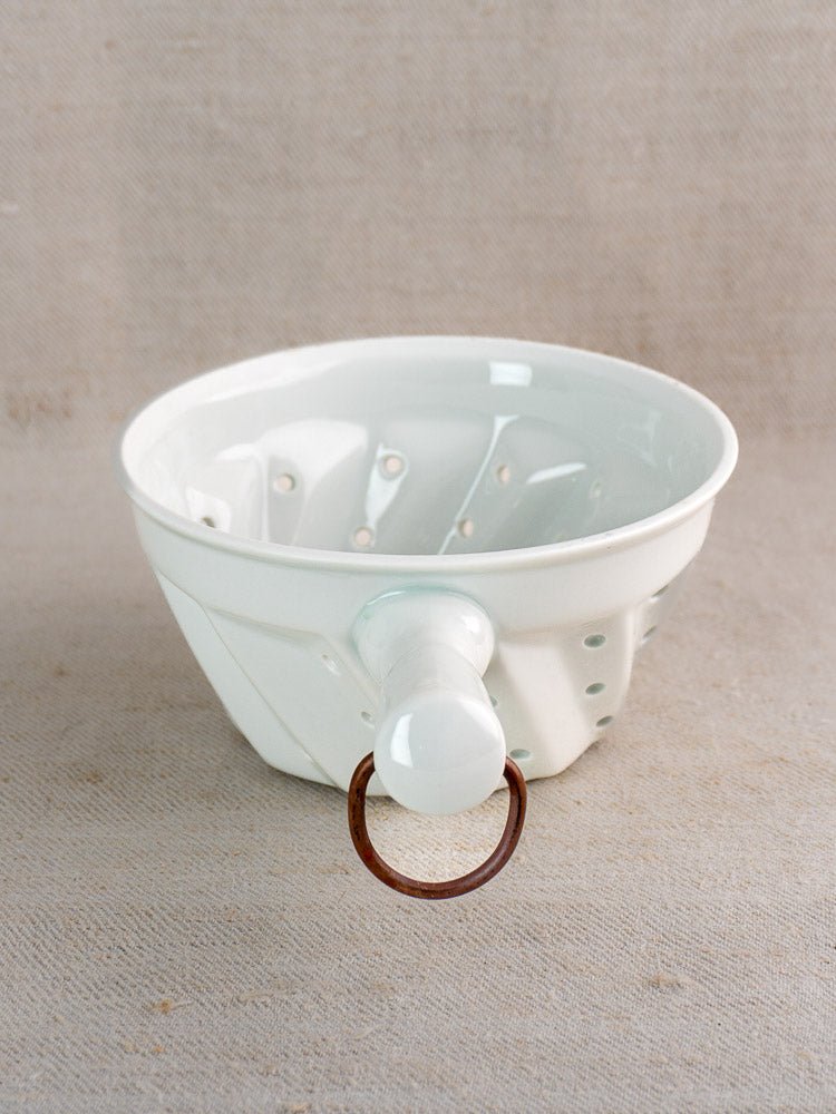 Vintage French porcelain berry strainer in white ceramic with handle and copper ring, on neutral surface.