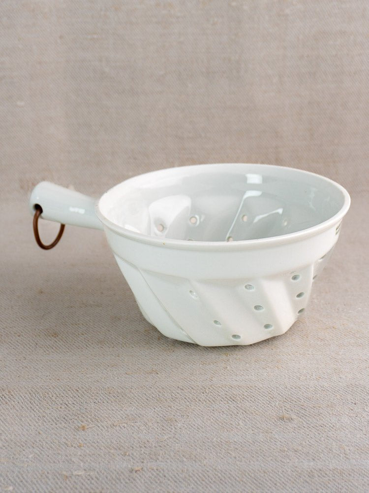 French porcelain colander with diagonal ridges, copper-accented handle, on neutral fabric background.