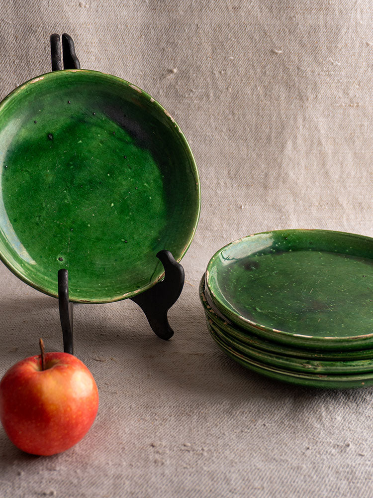 Set of five antique French green glazed earthenware plates, one upright on black stand, on textured beige fabric background.