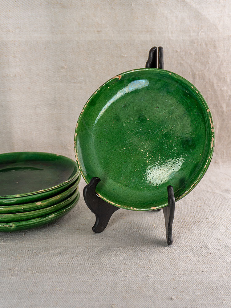 Set of five antique French green glazed earthenware plates, one on stand, four stacked, against textured beige fabric.