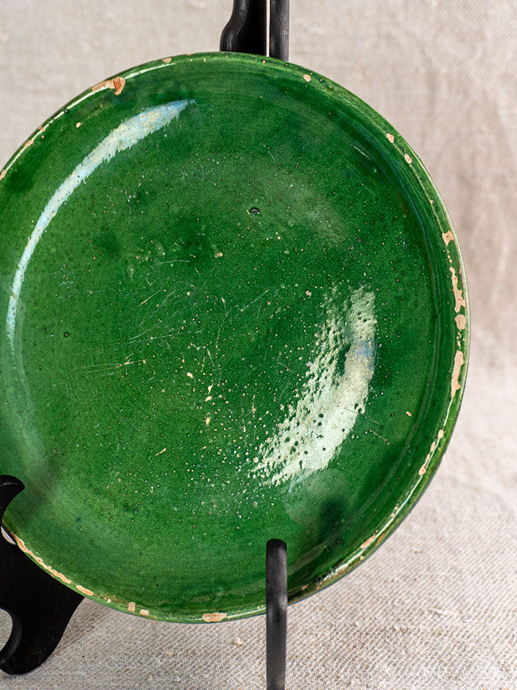 Set of five antique French green glazed earthenware plates with worn, chipped edges on a black stand against beige fabric.