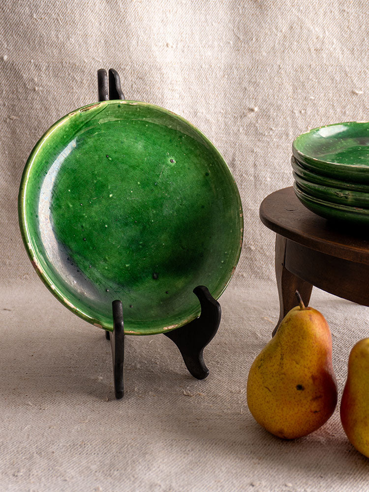 Antique French green glazed earthenware plate on black stand with stacked plates and two yellow pears on fabric.