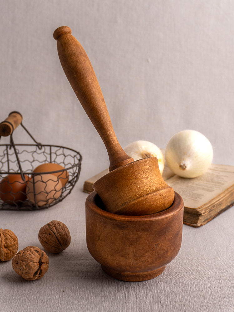 Antique French wooden mortar and pestle on light surface with walnuts, wire egg basket, onions, and open book.