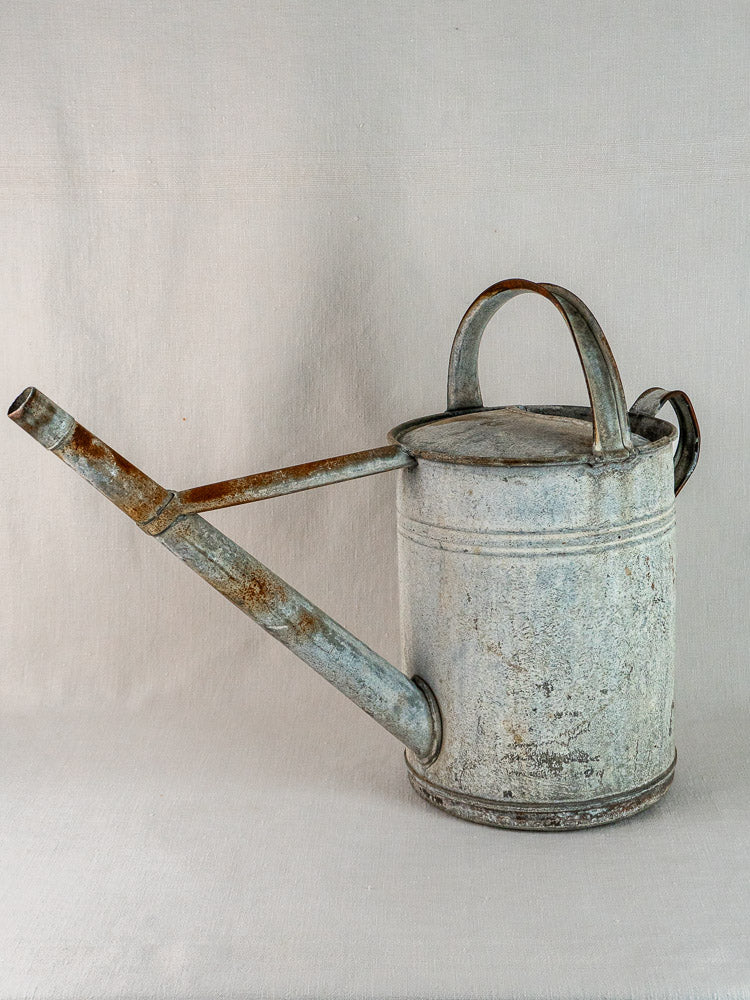 Weathered French zinc watering can with long spout and large handle on a neutral surface.