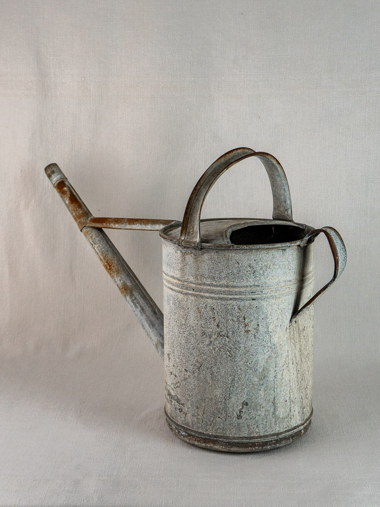 Vintage French-style zinc watering can with long spout and dual handles on neutral fabric.