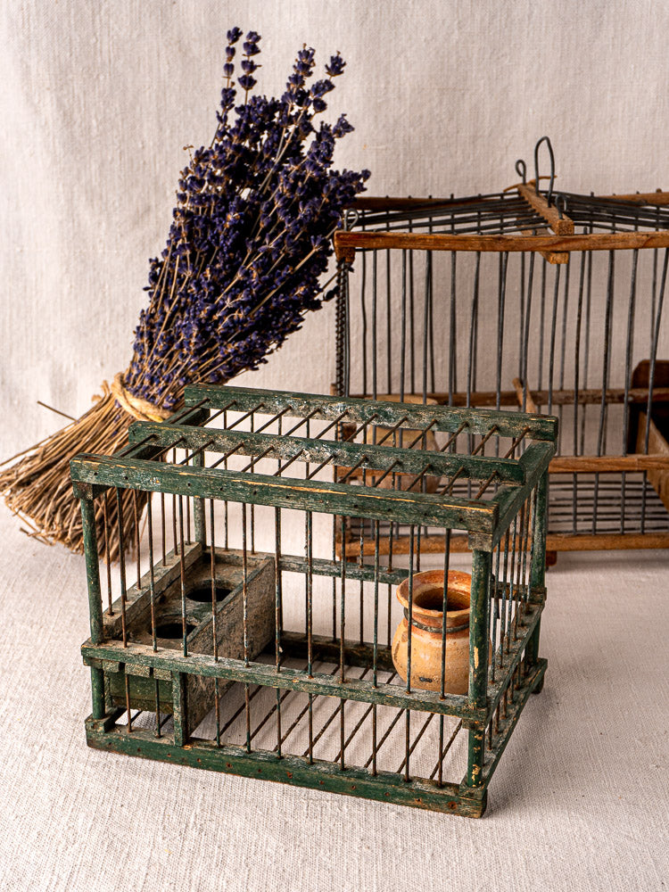 Antique French green painted wood and wire bird cage with terracotta cup, empty on neutral fabric background.