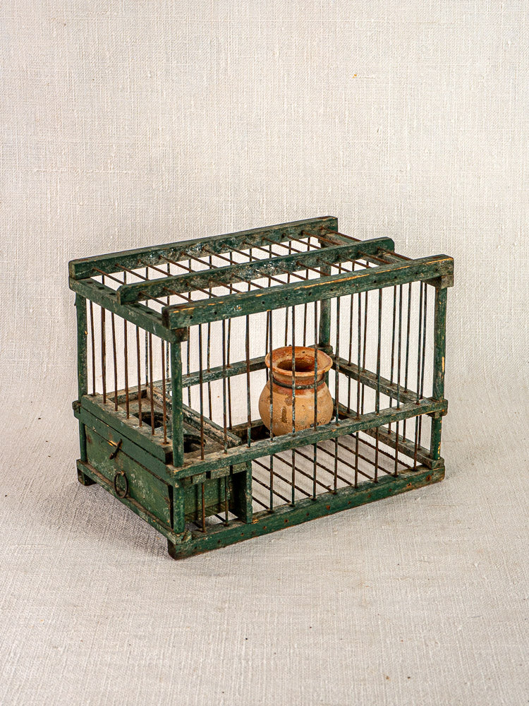 Antique French green painted wood and wire bird cage with terracotta cup, empty on neutral fabric background.