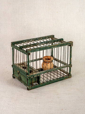 Antique French green painted wood and wire bird cage with terracotta cup, empty on neutral fabric background.