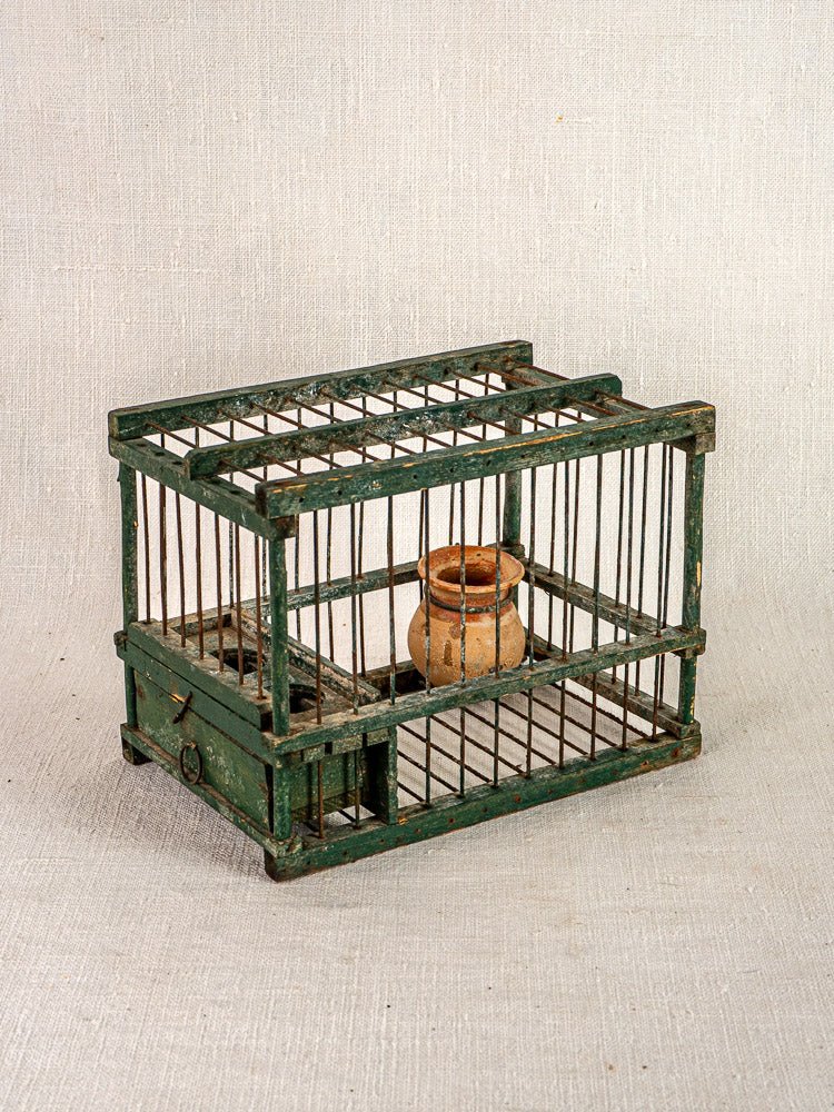 Antique French green painted wood and wire bird cage with terracotta cup, empty on neutral fabric background.