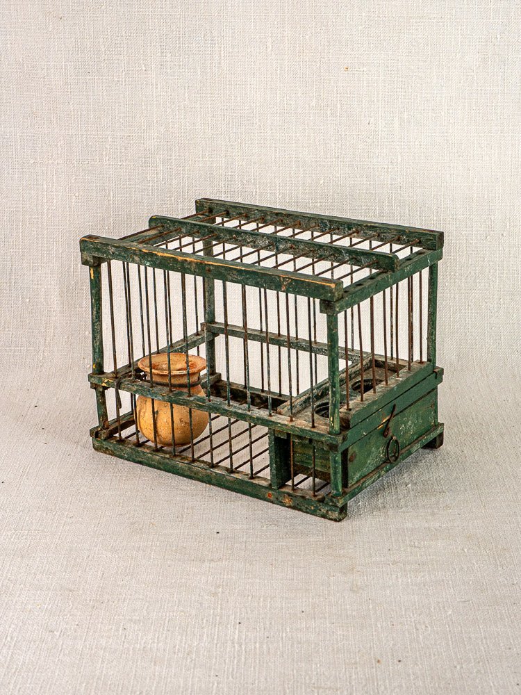 Antique French small wood and wire bird cage with vintage green paint, terracotta cup, on neutral fabric background.