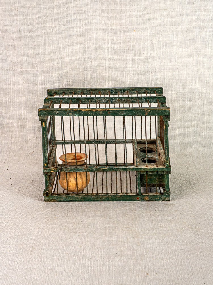 Antique French green painted wood and wire bird cage with vertical bars, terracotta cup inside, on neutral fabric background.