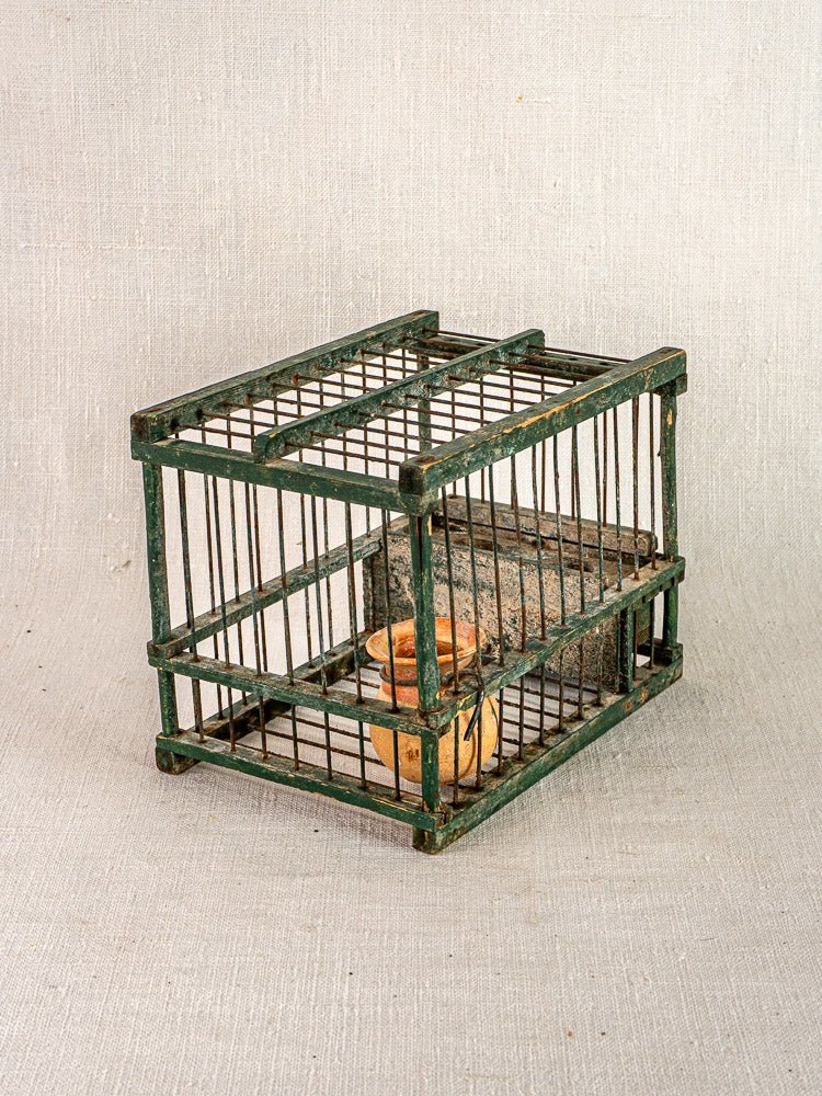 Antique French green painted bird cage with vertical wire bars and terracotta cup on neutral fabric background.