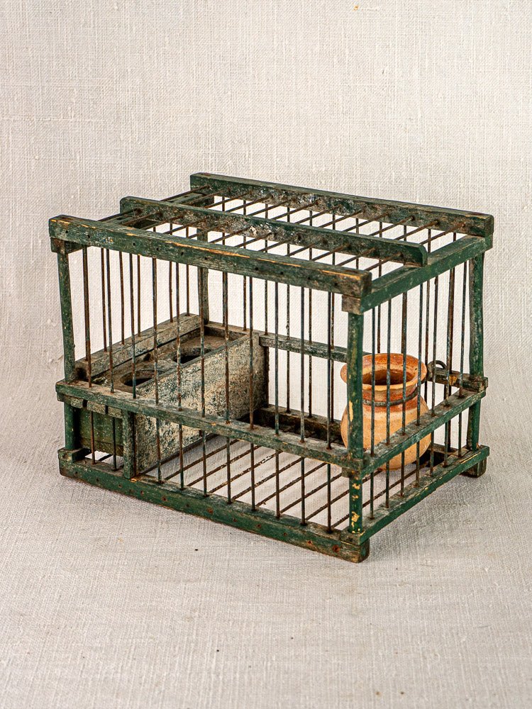 Antique French green painted bird cage with rustic wood, wire frame, and terracotta cup on neutral fabric background.