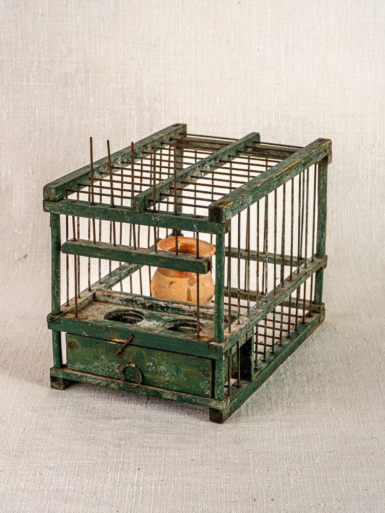 Antique French green painted wood and wire bird cage with terracotta cup, rusty bars, front lock on light textured surface.