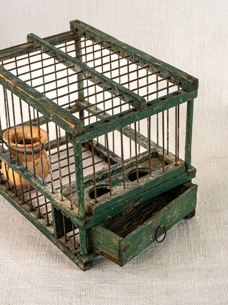 Antique French green painted wood and wire bird cage with terracotta cup and pull-out drawer on light fabric surface.