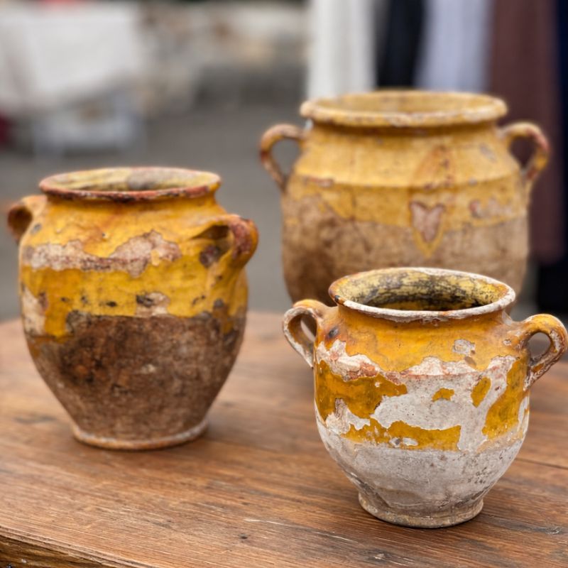 Three antique French confit pots with worn yellow glaze and exposed terracotta resting on a rustic wooden table